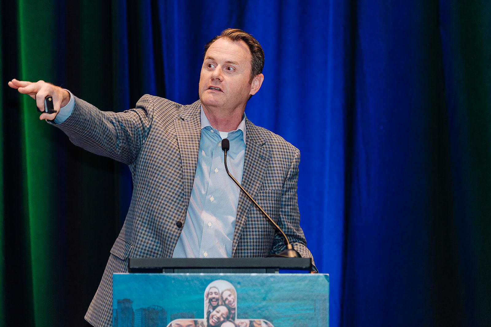 A corporate speaker presents from a podium beneath blue stage lighting